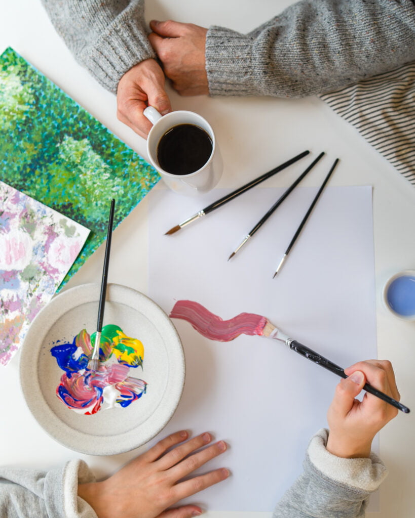 Kuva ylhäältä otettuna pöydän äärestä, missä näkyy aikuisen kädet pitelemässä kahvikuppia ja lapsen kädet maalamassa siveltimellä paperille.