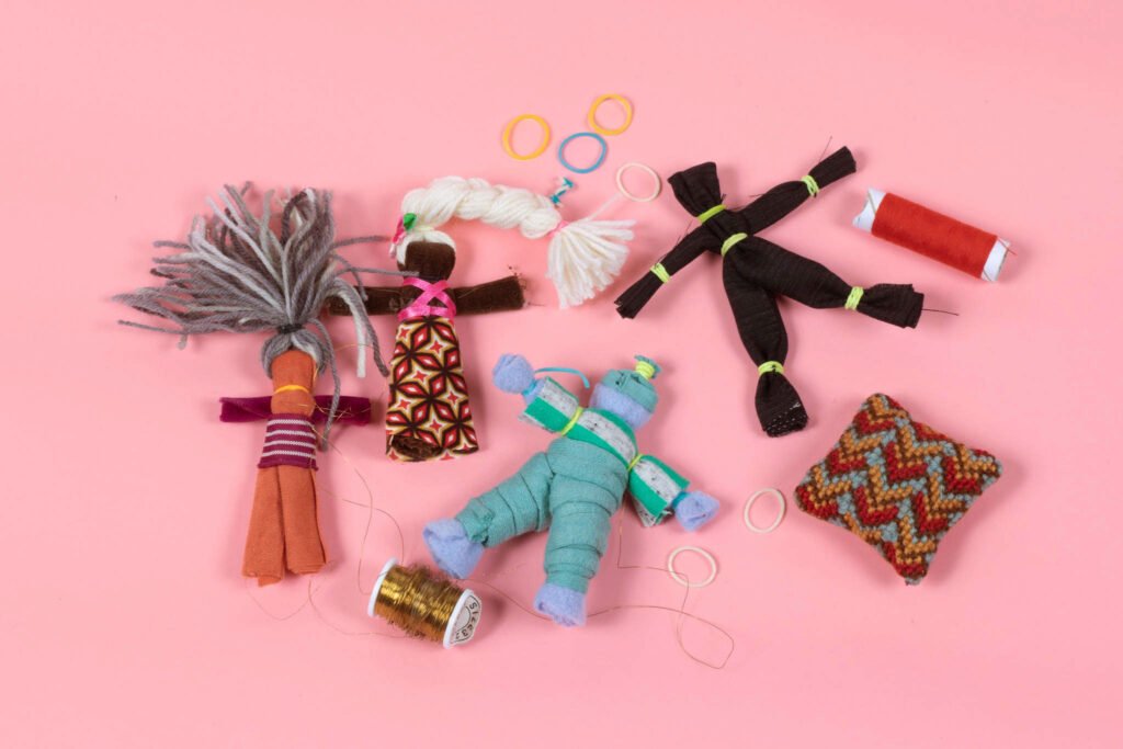 Kangasmateriaaleista valmistettuja pieniä erivärisiä nukkeja ja lankakeriä vaaleanpunaisella taustalla.