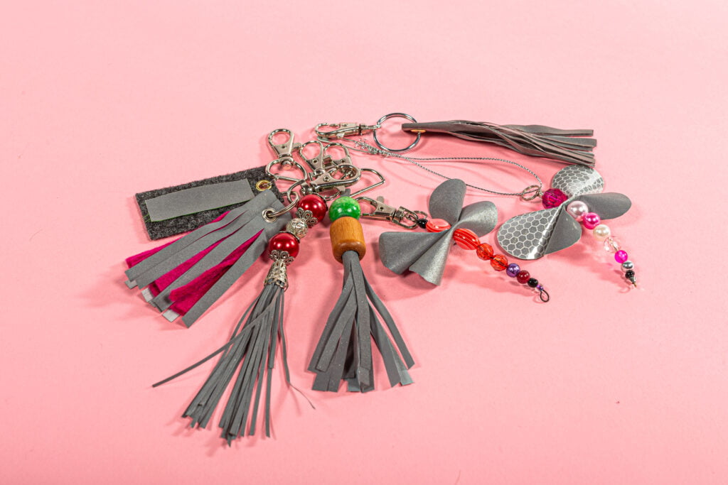 Seitsemän heijastinmateriaalista ja helmistä valmistettua avaimenperää vaaleanpunaisella taustalla.